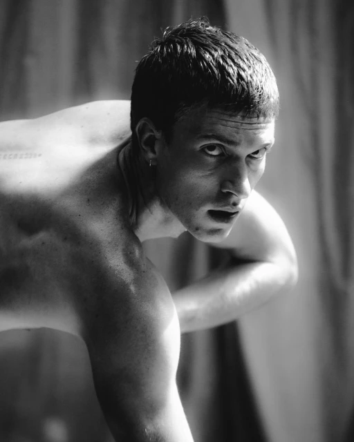 Black and white portrait of a shirtless adult male with short hair and an intense expression, posing in front of a textured backdrop.