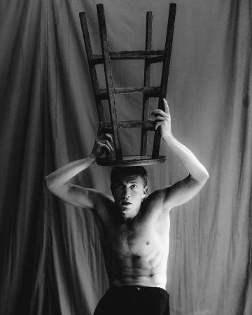 Black and white photo of a muscular man balancing a wooden chair on his head, posing shirtless against a draped background, lit from above.