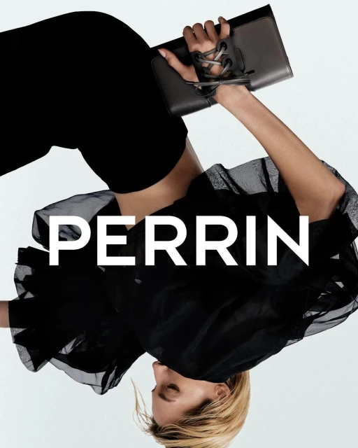 A woman models a black Perrin clutch. The model's dark outfit contrasts with the white background and highlights the accessory.