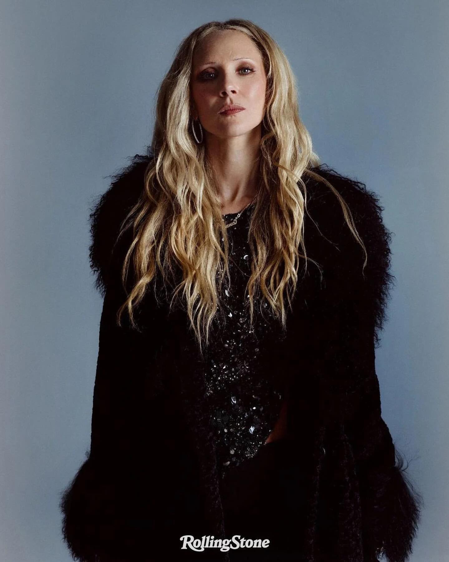 Portrait of a blonde woman wearing a black, feathered coat and sequined dress against a blue background, logo 'Rolling Stone' visible.