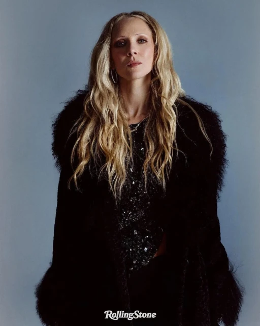 Portrait of a blonde woman wearing a black, feathered coat and sequined dress against a blue background, logo 'Rolling Stone' visible.