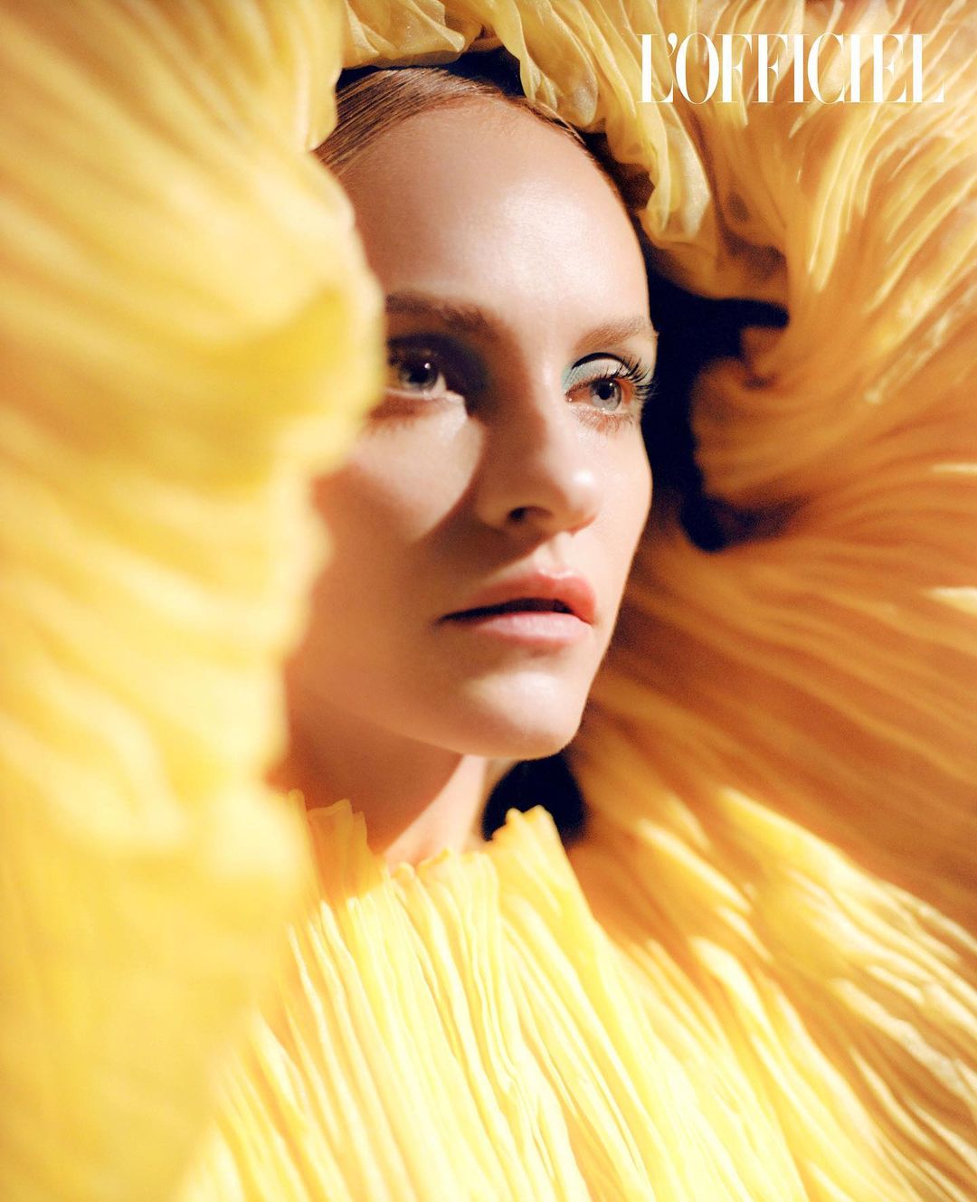 Close-up fashion shot of a fair-skinned woman with light hair and blue eye shadow surrounded by ruffled yellow fabric.