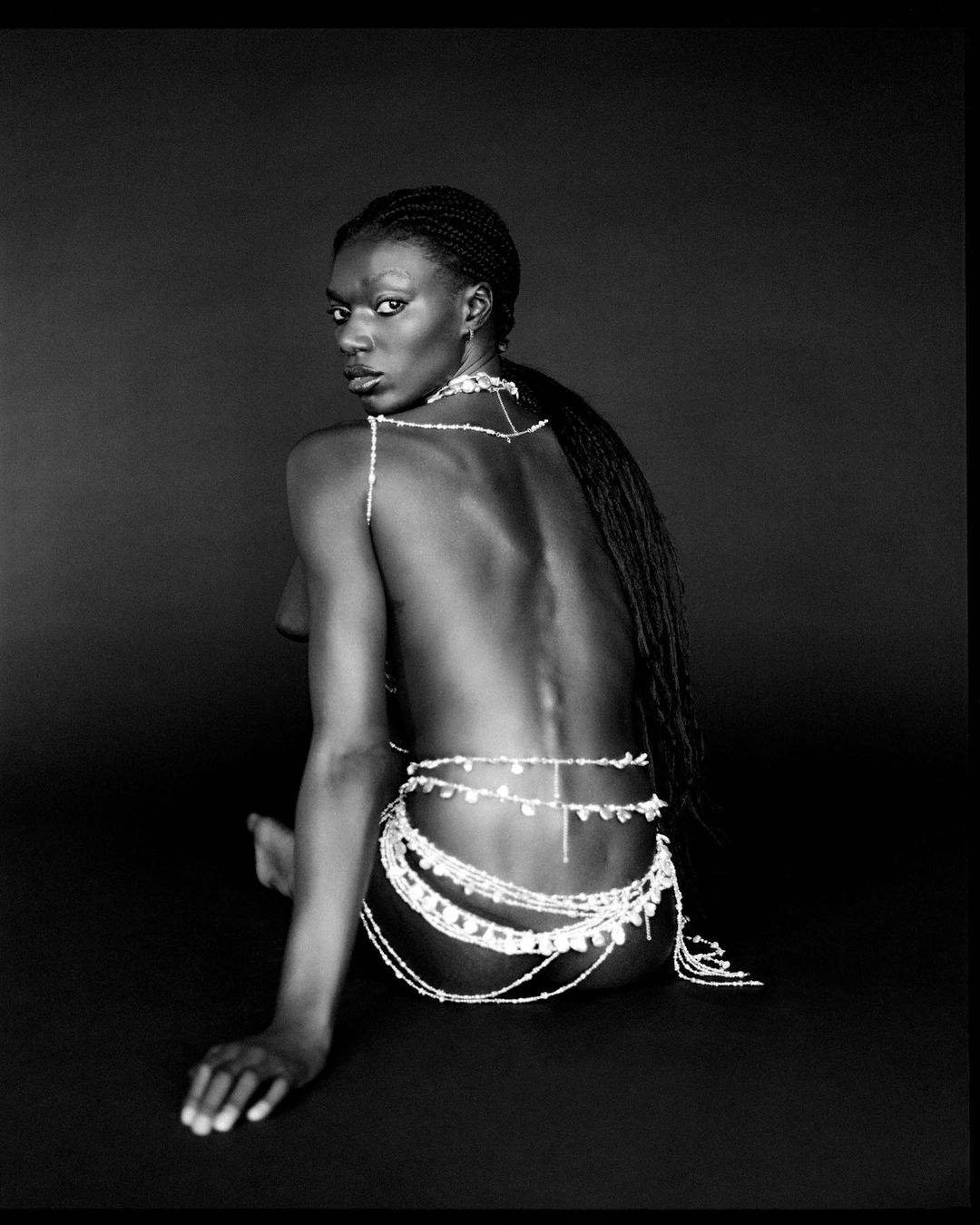 Black woman with braided hair wearing body jewellery seated against a dark background
