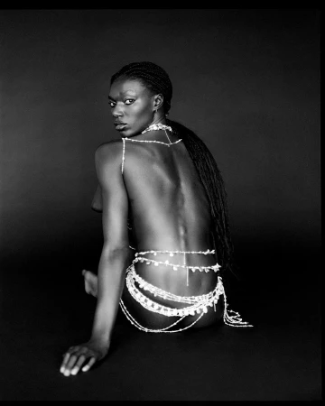 Black woman with braided hair wearing body jewellery seated against a dark background