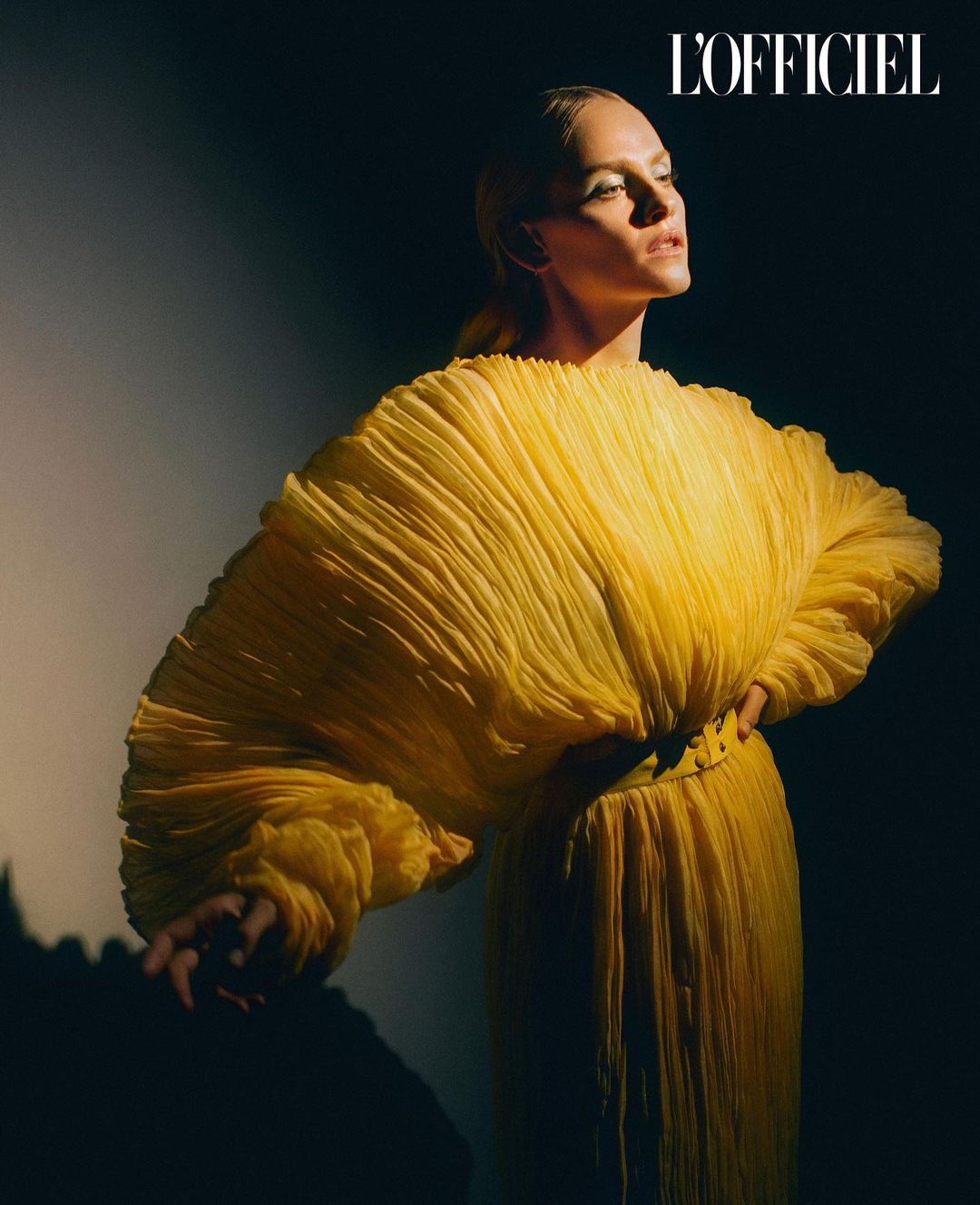 Elegant woman in a voluminous yellow dress poses dramatically against a dark background in a fashion editorial shot, looking away.