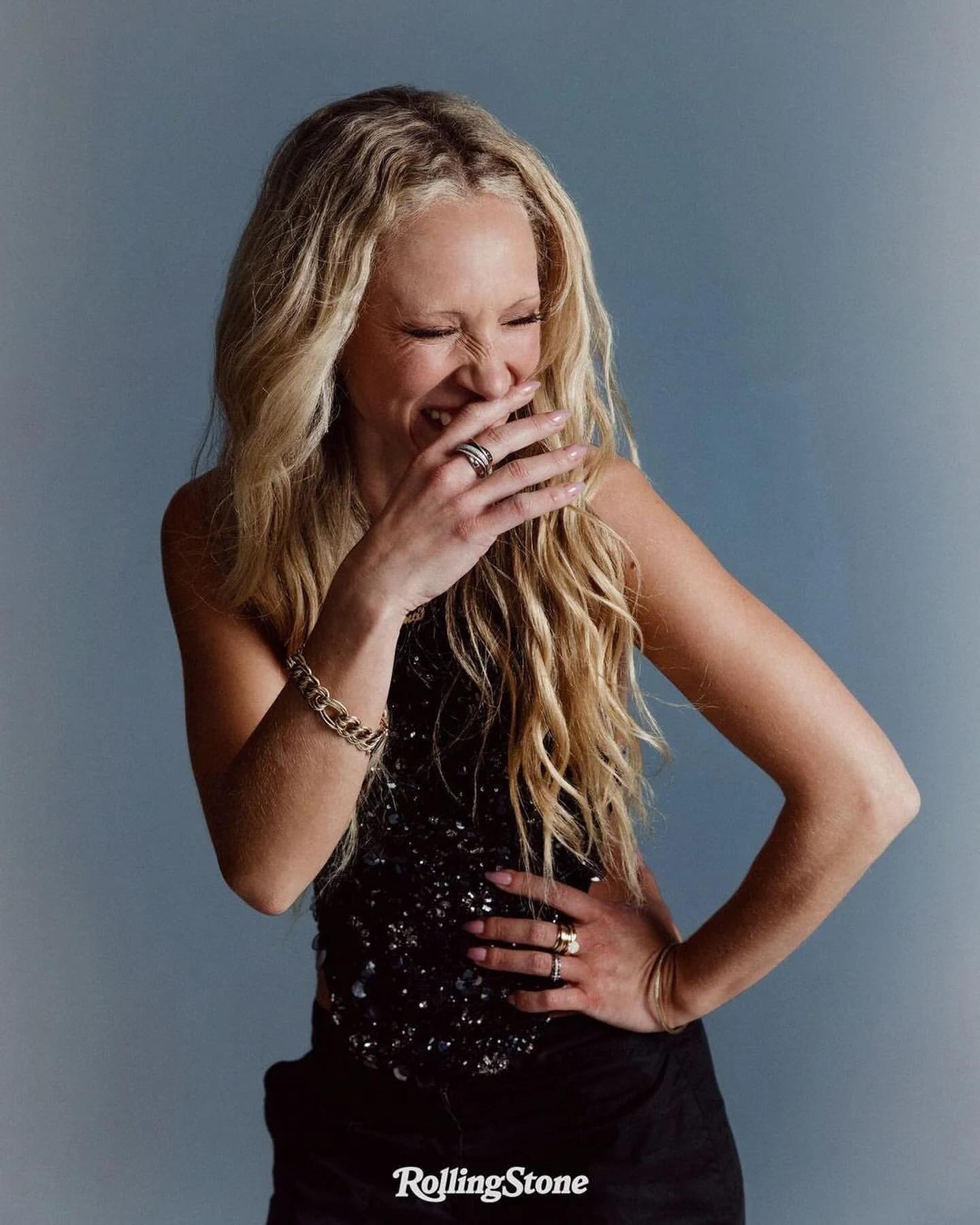Blonde woman laughing with hand covering mouth wearing a dark sequined top against a grey backdrop photographed by Yana Yatsuk