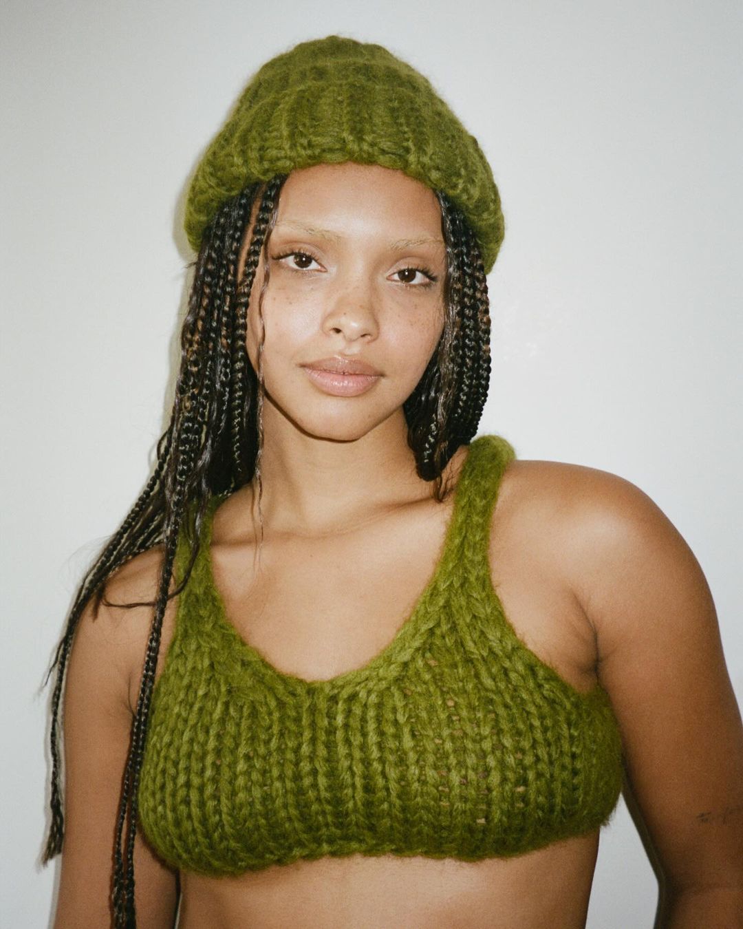 A young adult female with braided hair is wearing a green knit beanie and top in a studio shot against a plain white background.