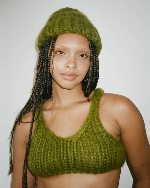 A young adult female with braided hair is wearing a green knit beanie and top in a studio shot against a plain white background.