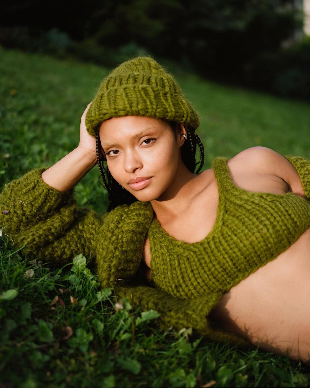 Woman in green knitted hat and matching top, laying in a grassy field with soft sunlight, in a fashion or lifestyle setting.
