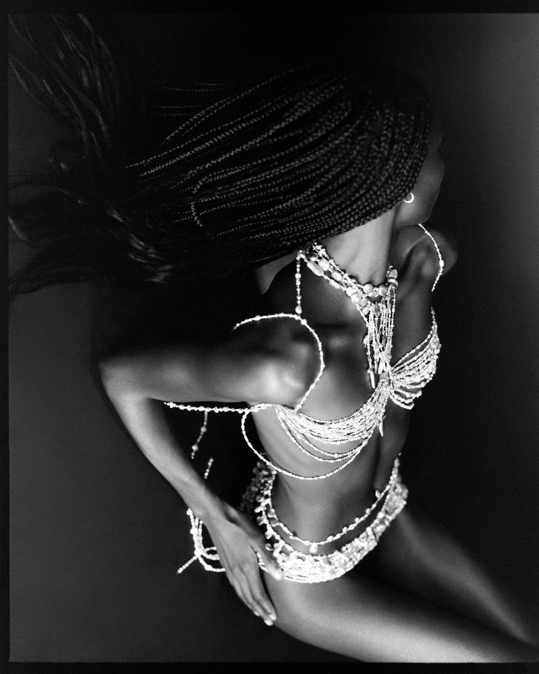 Black and white studio shot of an adult female with braided hair wearing beaded jewellery, posing overhead with a dark background.