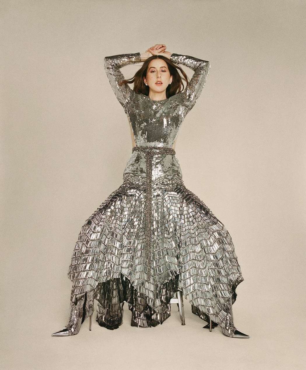 A woman with dark hair poses indoors wearing a long-sleeved silver sequin dress and matching heels on a cream-colored seamless background.