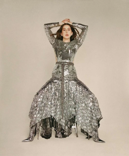 A woman with dark hair poses indoors wearing a long-sleeved silver sequin dress and matching heels on a cream-colored seamless background.