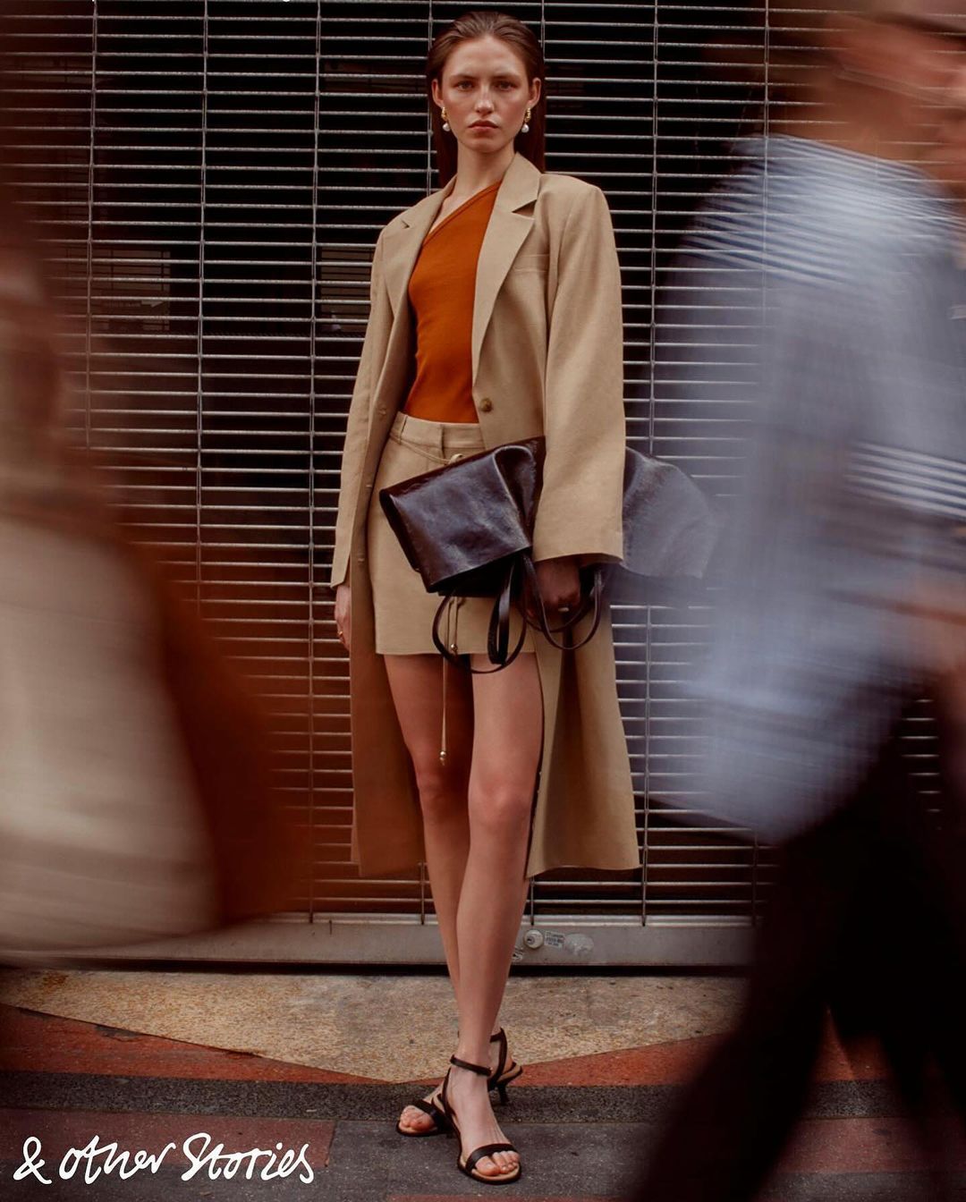 Fashion photo of a woman in a tan skirt and jacket, orange top, and black sandals, holding a black bag in front of a metal gate.