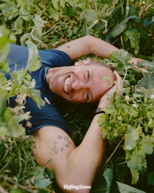 Smiling adult male lying in green foliage outdoors with arm raised, wearing a blue t-shirt and wrist watch, sunny day, portrait