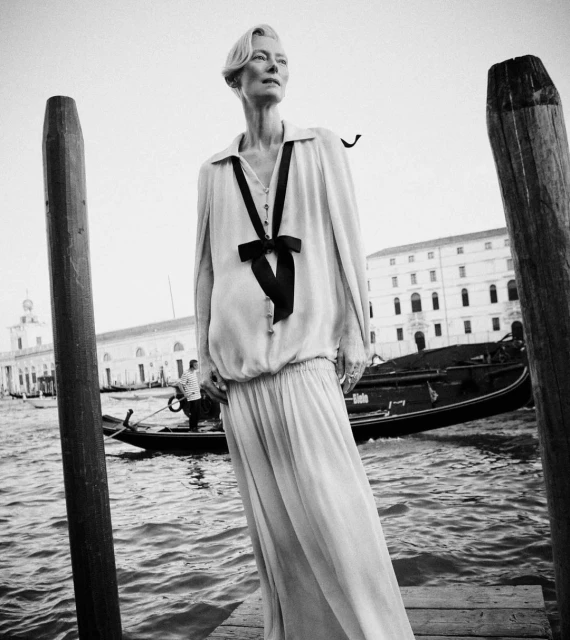Black and white portrait of an elegant woman in a long dress with a bow, standing by the water near wooden posts and a gondola.