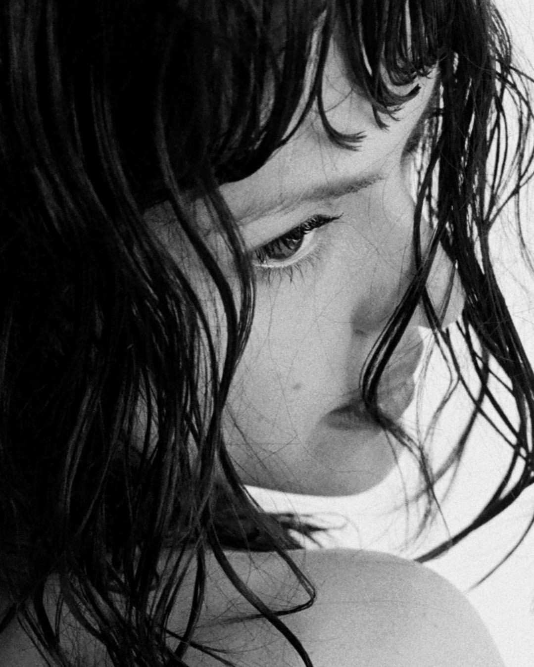 Monochrome portrait of a young person with wet, dark hair framing their face, looking down and to the side with a contemplative expression.