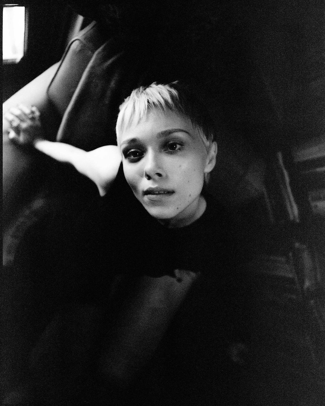 Monochrome portrait of a fair-skinned woman with short blonde hair looking up with a neutral facial expression in a dark indoor setting.