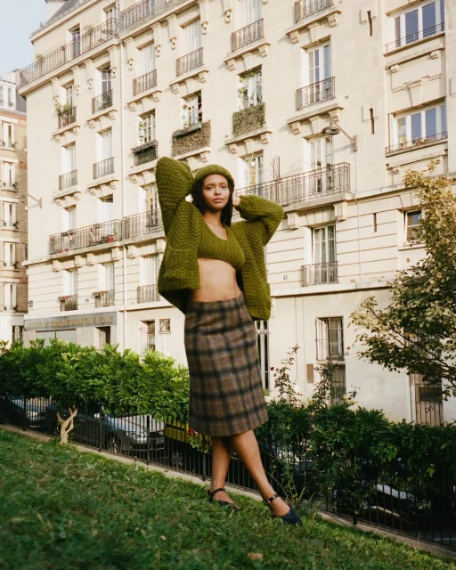 A young woman poses outdoors in a green knit top and cardigan with a brown plaid skirt against a building and green foliage.