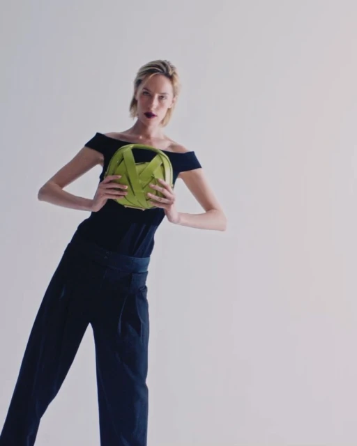 Blonde woman in black off-shoulder top and trousers holding a green woven handbag, posing in a bright studio.