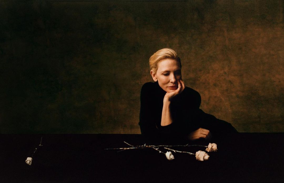 Portrait of an adult woman in a black turtleneck with short blonde hair, posing with white roses in a dark, moody studio setting.