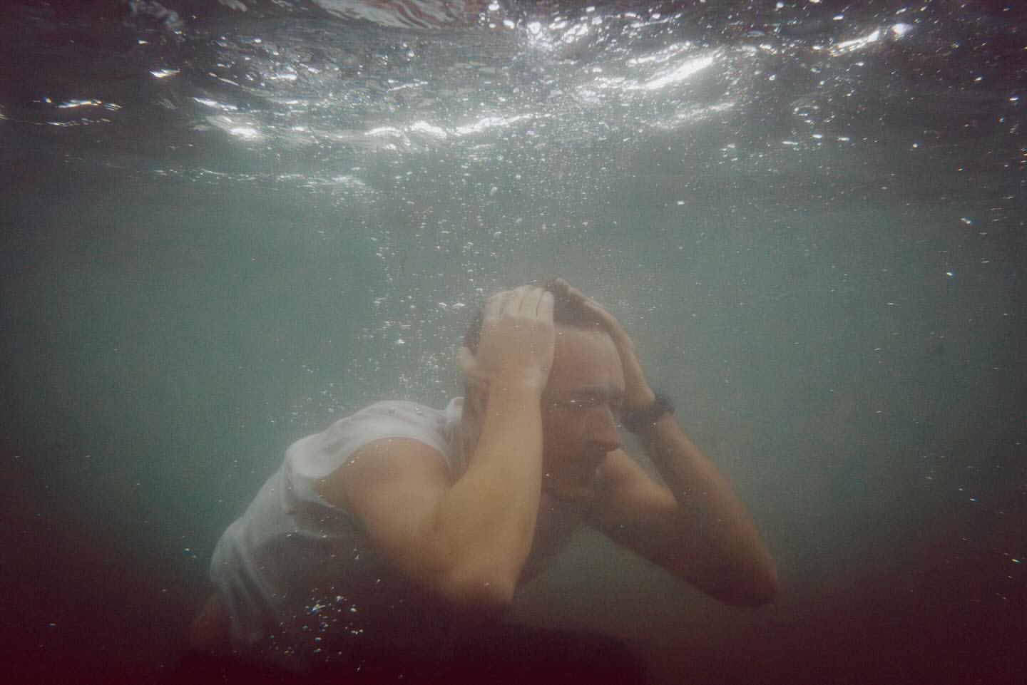 Underwater portrait of a man in a white shirt with his hands on his head, surrounded by bubbles and diffused natural lighting.