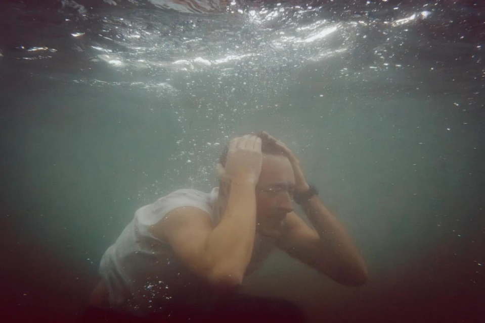 Underwater portrait of a man in a white shirt with his hands on his head, surrounded by bubbles and diffused natural lighting.