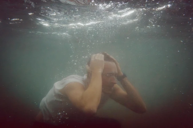 Underwater portrait of a man in a white shirt with his hands on his head, surrounded by bubbles and diffused natural lighting.