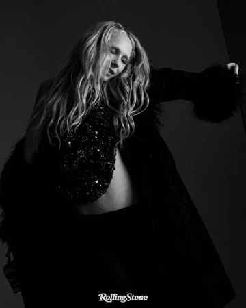 Portrait of a blonde woman wearing a black embellished top and dark coat against a dark background