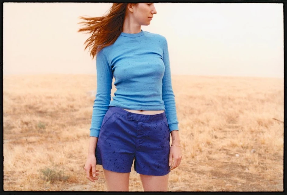 Young adult woman stands in an open field wearing a light blue long-sleeved top and dark blue shorts, with wind blowing her red hair.