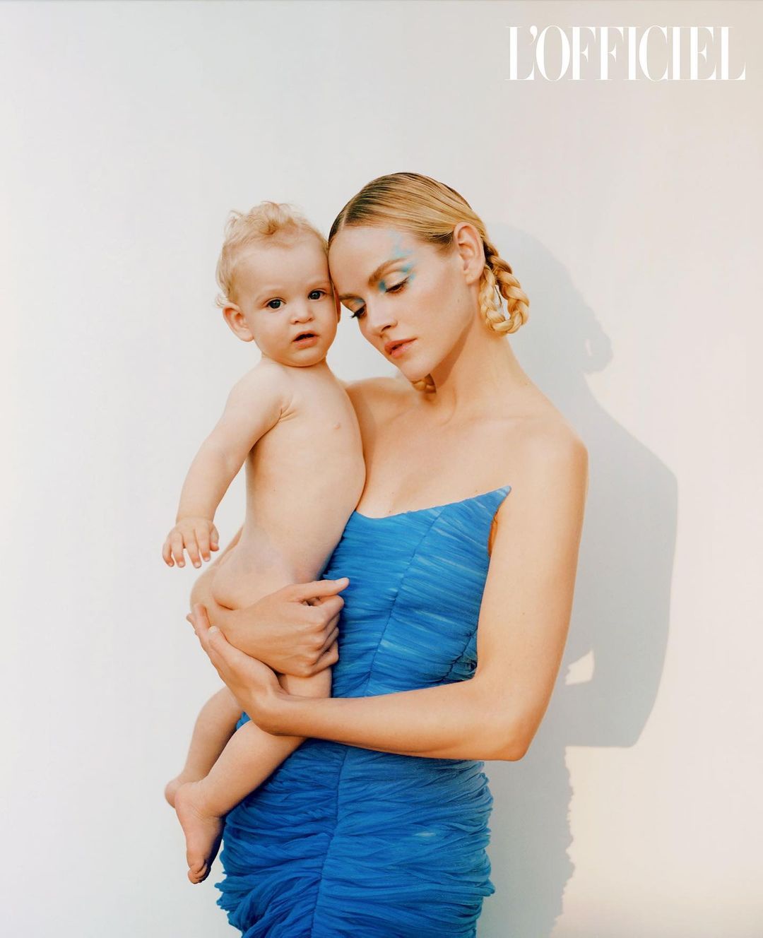 Portrait of a mother holding her baby, both with fair skin, in front of a neutral wall. The mother wears a blue dress with makeup.