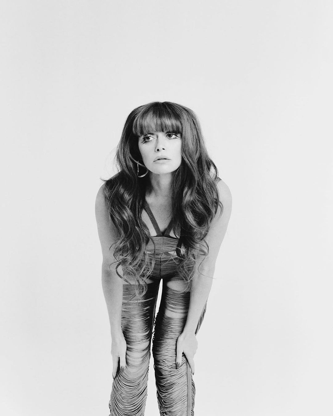 Monochrome portrait of a woman with long wavy hair and bangs, leaning forward, wearing ripped jeans against a plain backdrop.