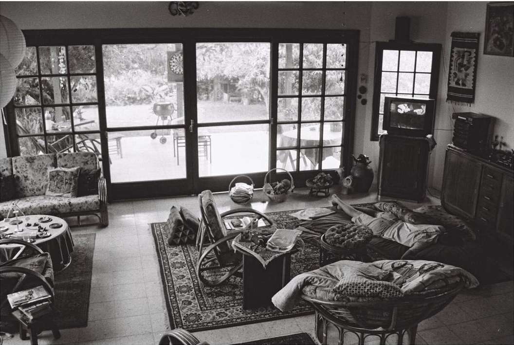Black and white indoor scene features a woman relaxing on cushions near a television, with doors opening to a garden.