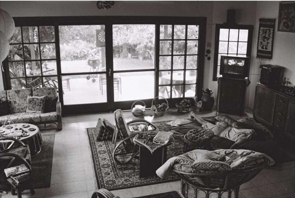 Black and white indoor scene features a woman relaxing on cushions near a television, with doors opening to a garden.