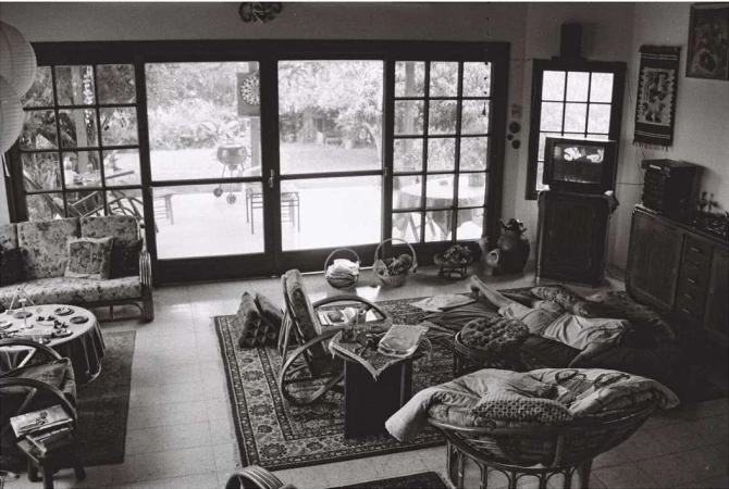 Black and white indoor scene features a woman relaxing on cushions near a television, with doors opening to a garden.