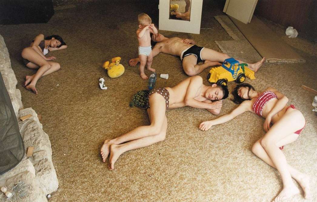 Several people and a toddler lie on the floor in a home interior with a fireplace and toys, bathed in soft daylight. 