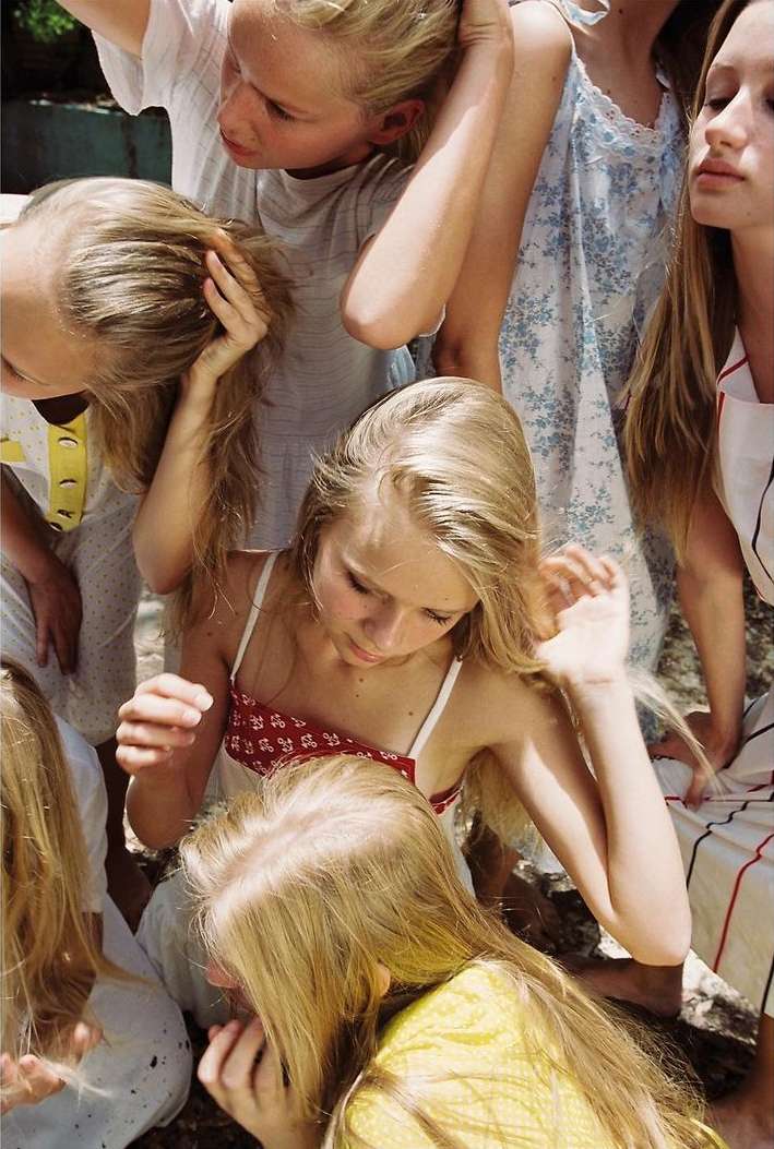 Group of young girls with long blonde hair in various poses and outfits, possibly pajamas, in an outdoor setting with diffused light.