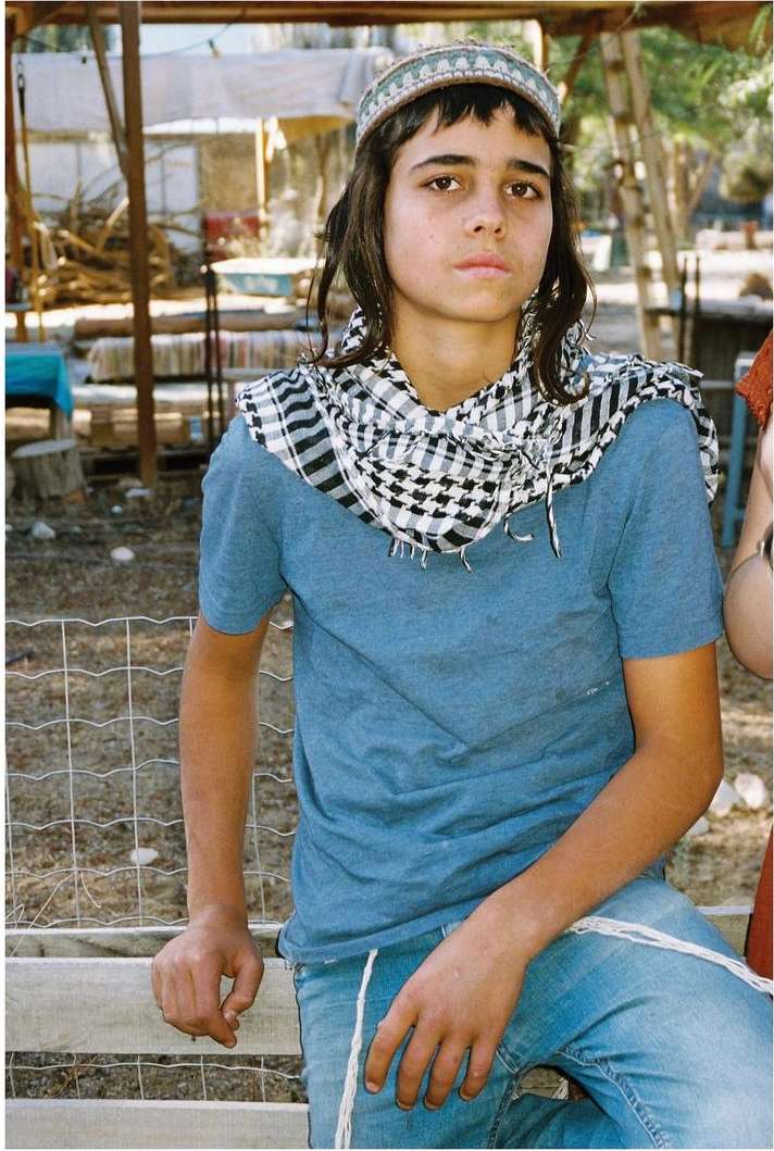 Teenager with dark hair and head covering wearing a blue t-shirt and jeans, sitting outdoors near a fence, photographed at eye-level.