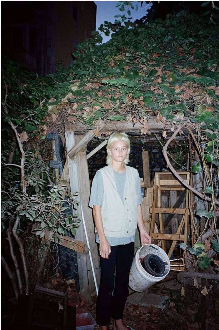 Young adult carrying a bucket outdoors amid green foliage, in front of a wooden structure in a yard setting, captured in natural light.