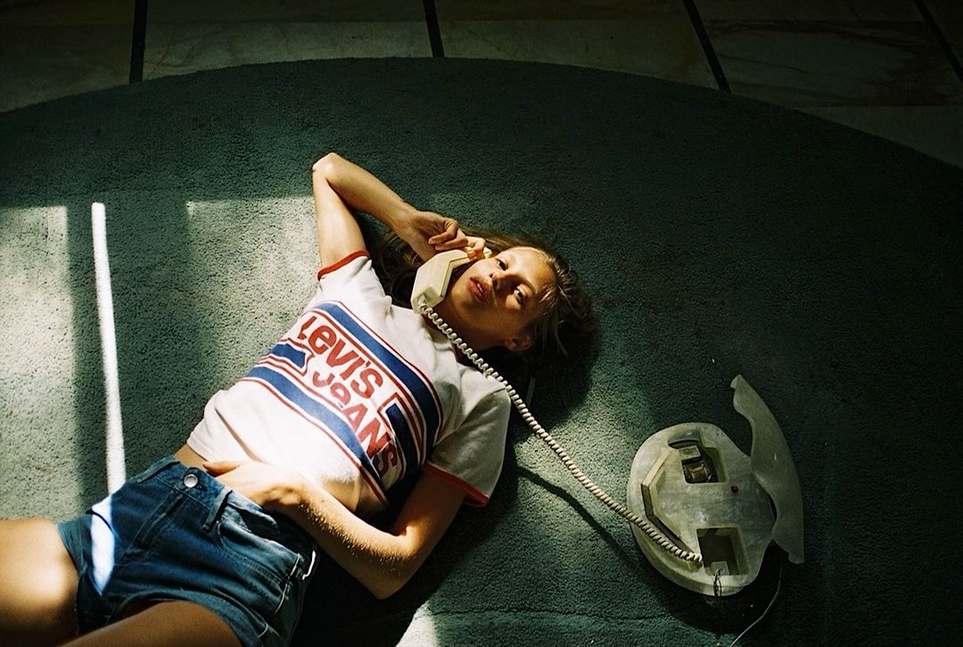 A young adult woman with brown hair wearing a Levi's T-shirt and denim shorts is lying on a green carpet and talking on a vintage phone.