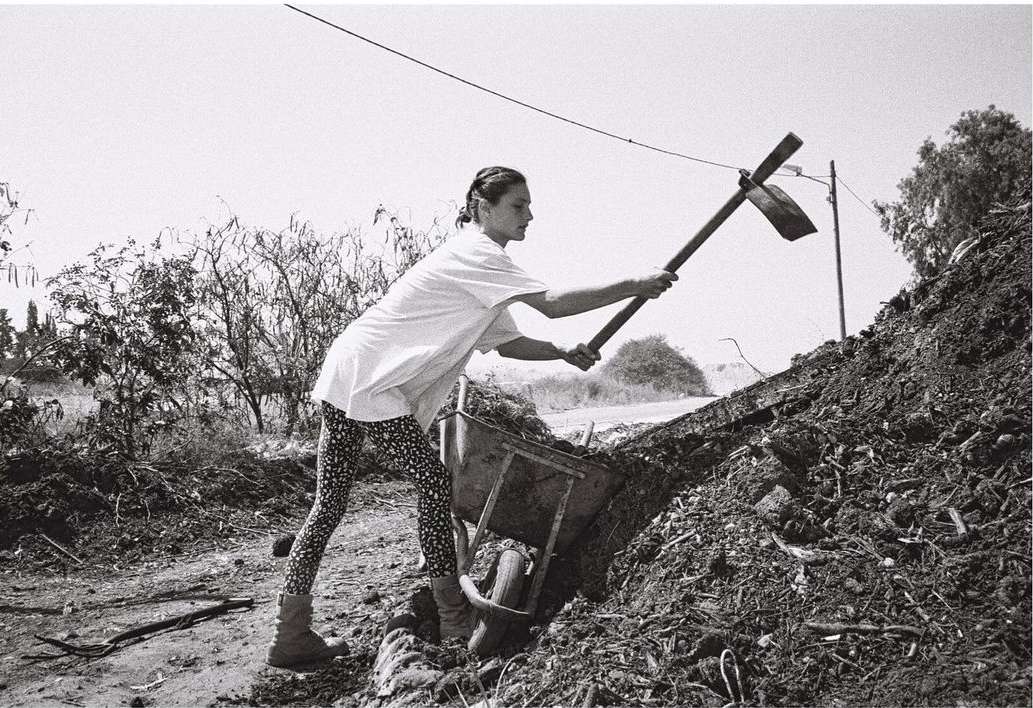 Black and white photograph of an adult female farm worker digging into a pile of dirt with a pickaxe, wearing leggings and boots.