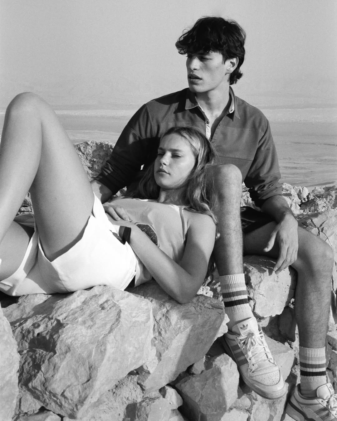 Black and white portrait of a young adult couple lounging on rocks by the ocean. Woman laying on rocks with man seated behind her.