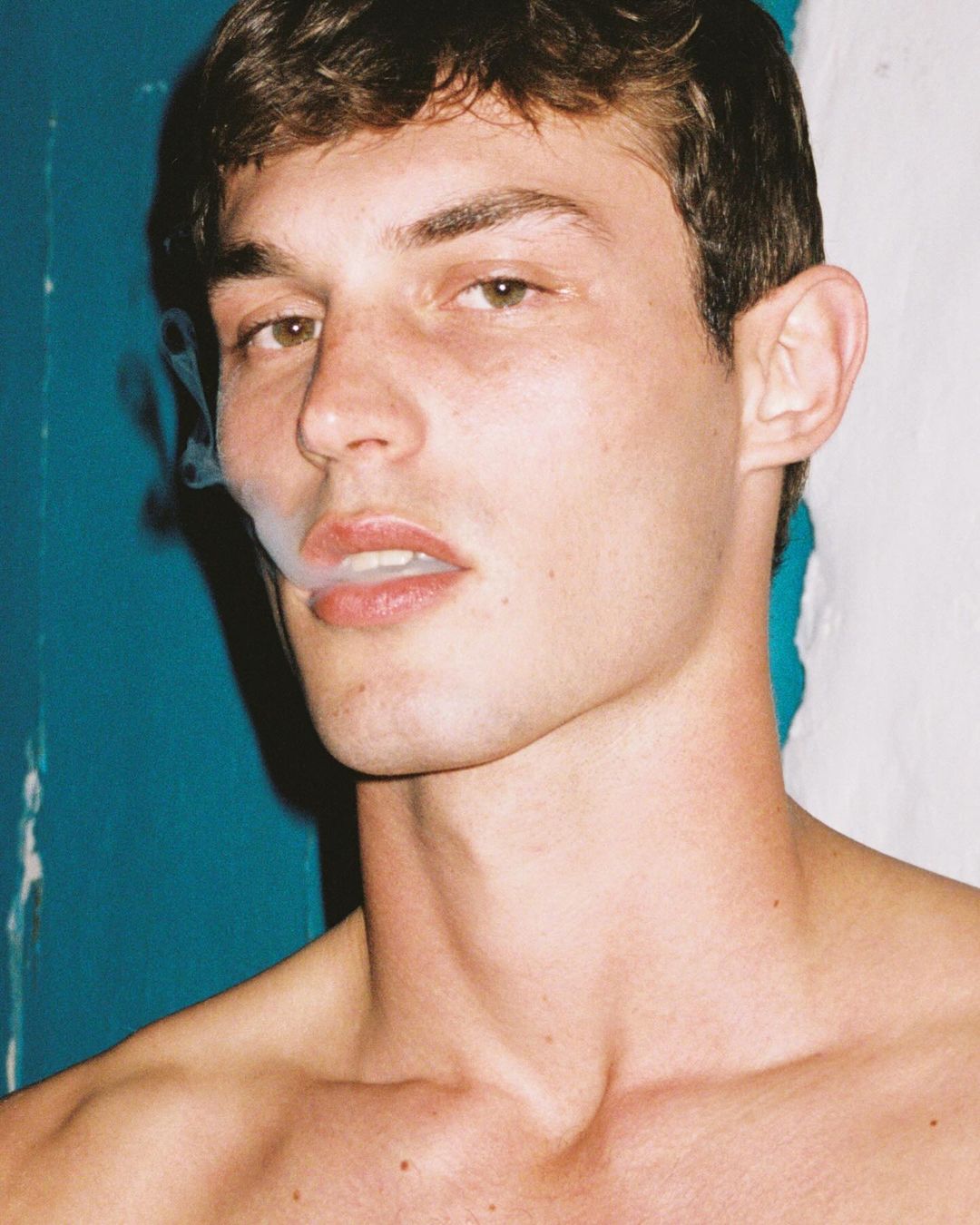 Close-up of a young adult male with short brown hair, gazing toward the viewer while exhaling smoke, set against a split blue and white background.