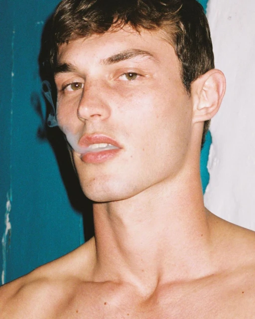 Close-up of a young adult male with short brown hair, gazing toward the viewer while exhaling smoke, set against a split blue and white background.