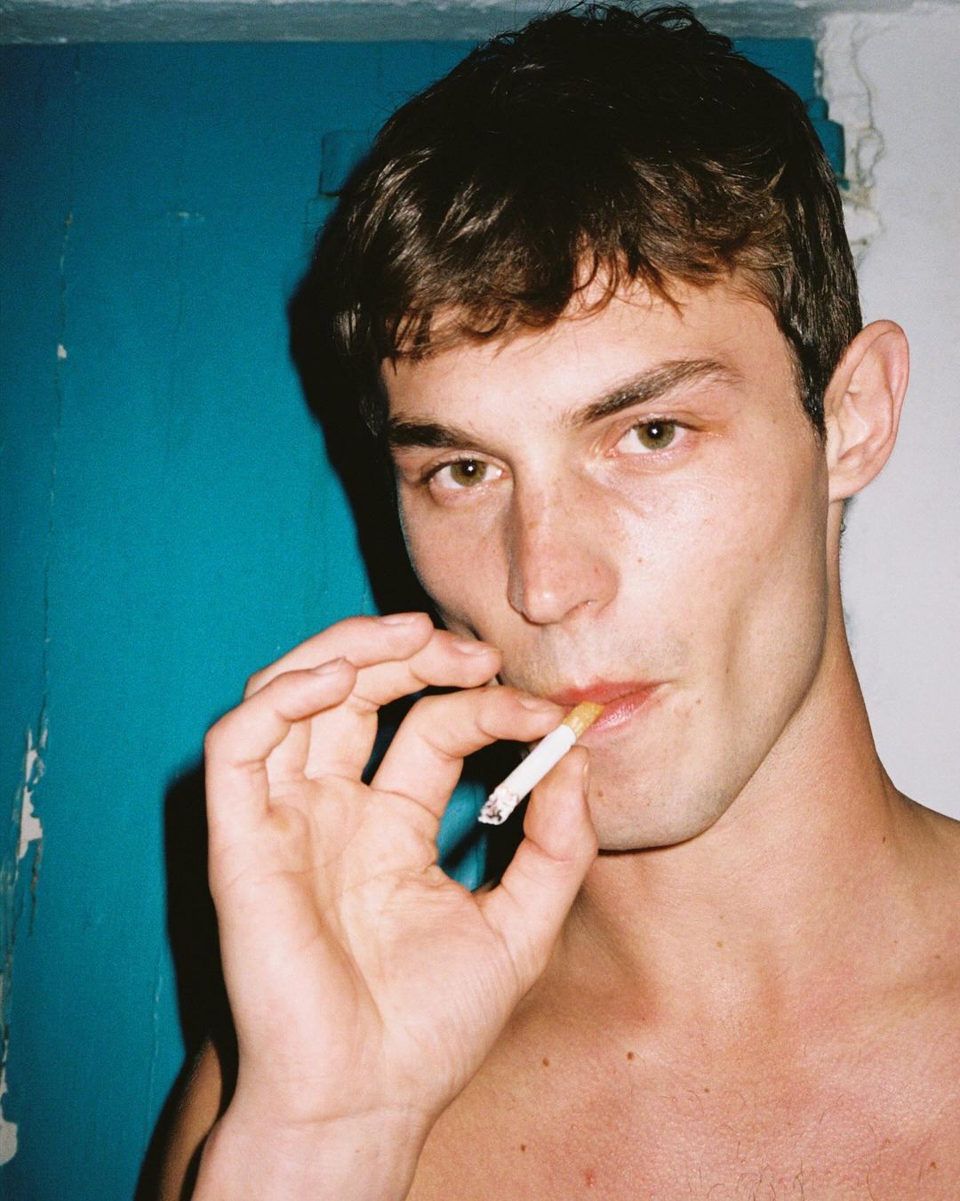 Portrait of a young adult male indoors against a blue wall, smoking a cigarette, captured in natural lighting with a close-up shot.