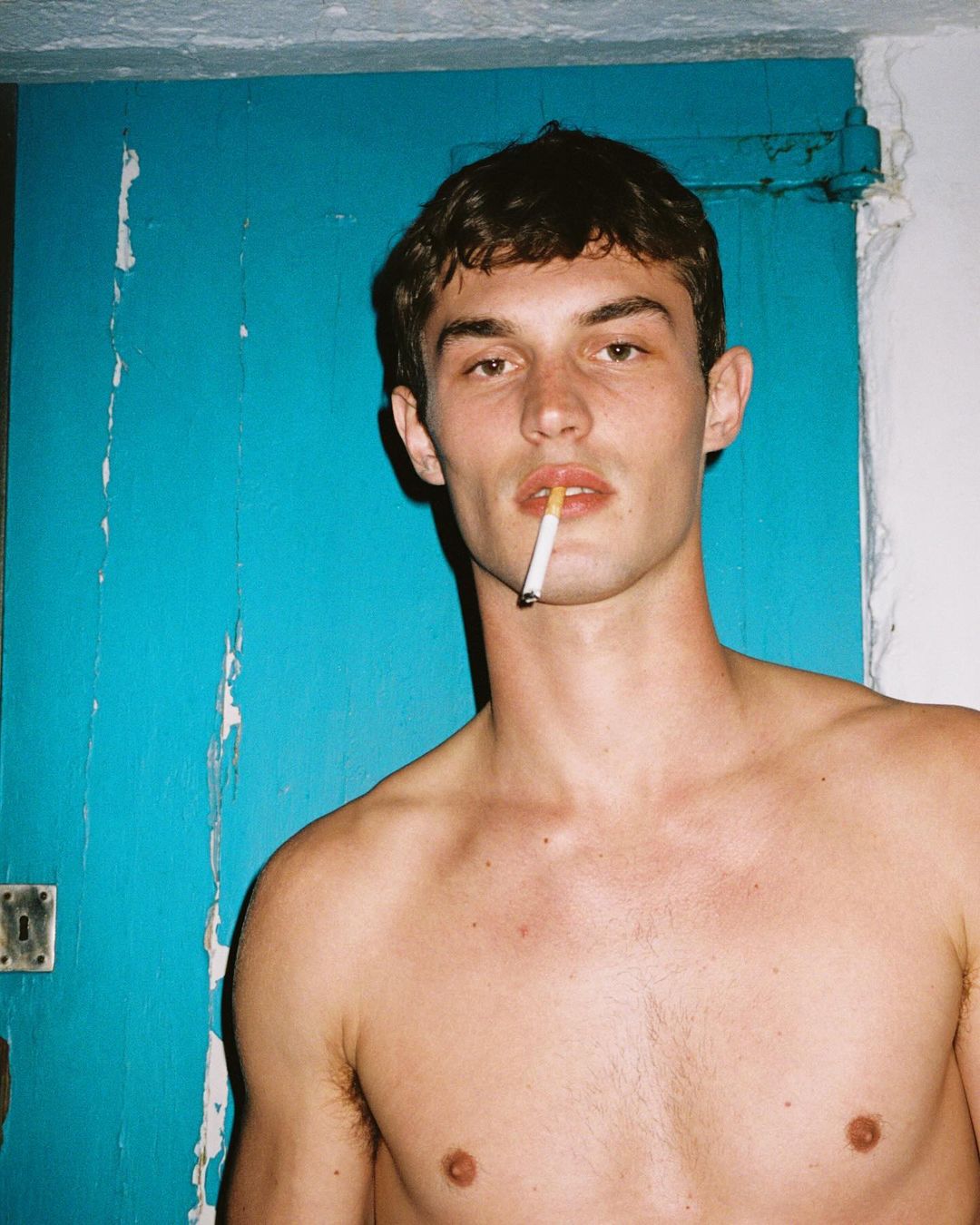 Portrait of a shirtless young man with a cigarette in his mouth standing in front of a blue door, captured in soft, natural light.