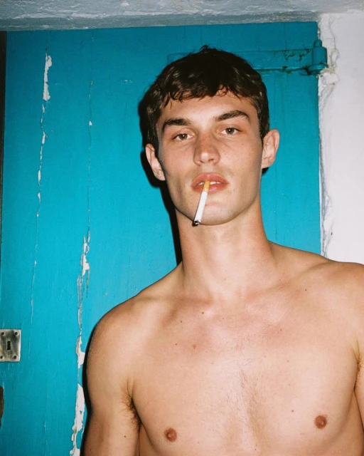 Portrait of a shirtless young man with a cigarette in his mouth standing in front of a blue door, captured in soft, natural light.