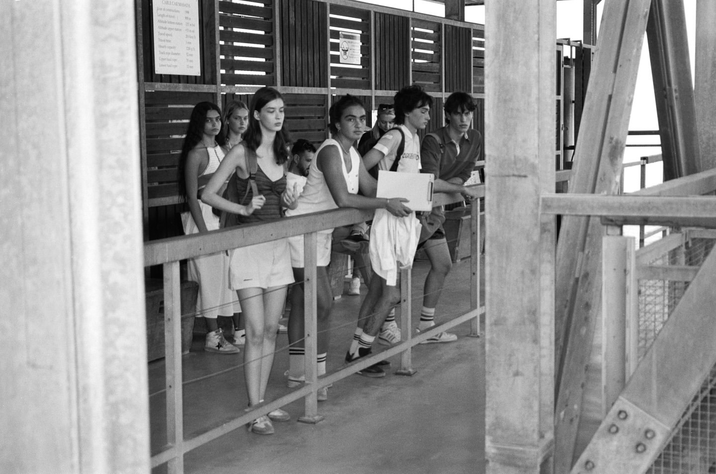 Black and white shot of a group of young people standing and leaning against a railing, looking out at an industrial structure.