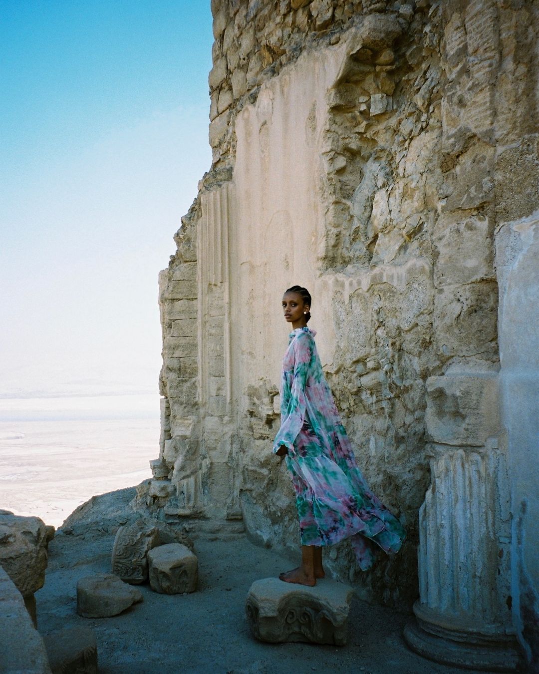 A fashion shot of a barefoot woman in a long floral dress, standing on a rock amid the ruins of an ancient stone structure.