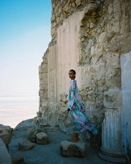 A fashion shot of a barefoot woman in a long floral dress, standing on a rock amid the ruins of an ancient stone structure.