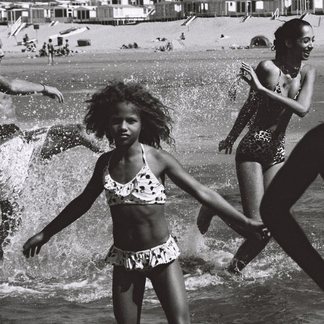 Black and white photograph of a child in a polka-dot bikini walking through the water on a beach with adults and distant beach houses.